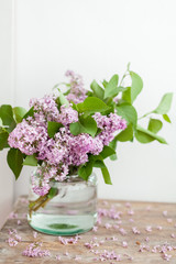 Lilac flowers bouquet with greenery branches in a glass vase on the wooden coffee table. Flower mock up vertical orientation