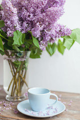 Obraz na płótnie Canvas Empty light blue coffee cup on the table. Violet lilac flowers bouquet in glass vase on the wooden coffee table. Kitchen interior decorations. Mother's day congratulation