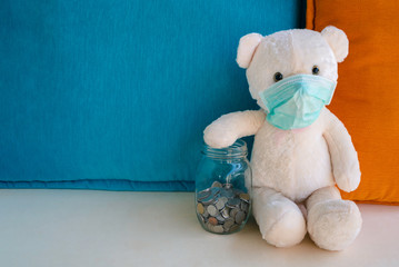 Teddy bear with surgical face mask holding glass bottle money savings with coins in hand worry about illness, insufficient money to pay medical expenses. Health insurance, medicare and finance concept