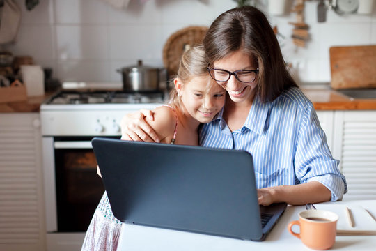 Working from home office with kid. Happy mother and daughter shopping online, using laptop together. Woman hugging child. Freelancer workplace at kitchen table. Female business, virtual communication.