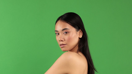 Portrait of beautiful young asian girl on green background with bare shoulder turned to camera and looking at camera