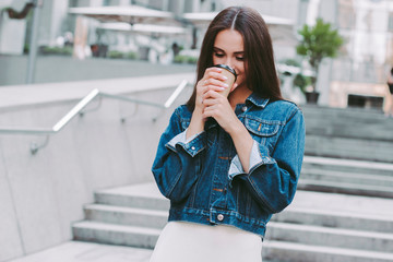 Young happy hipster girl in stylish denim jacket enjoying morning coffee on the go. Beautiful...