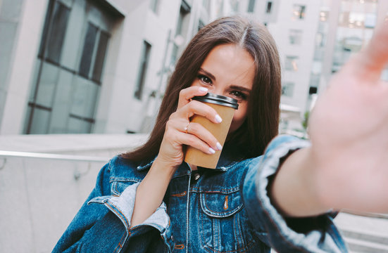 Young beautiful woman in denim jacket taking selfie on camera while drinking takeaway coffee outdoors. Cheerful attractive hipster girl making photo on phone with coffee cup in hand. Coffee on the go