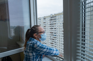 A woman in a medical mask at home quarantine coronavirus on the balcony, a view of the city of multi-storey buildings. The concept of self isolation stay at home