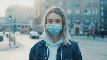 Young woman in medical mask standing in city street looking to camera road with cars on background Concept of health and safety life COVID-19 coronavirus virus protection pandemic in world