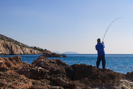Unrecognizable fisherman a man stands on the sea shore with rocks and pulls the rod, biting the fish. - Powered by Adobe