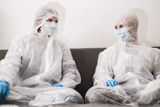 Two Person In Protective White Translucent Suit, Blue Rubber Gloves, Medical Mask Are Sitting On Sofa At Home. Stay Home During The Coronavirus Pandemic.