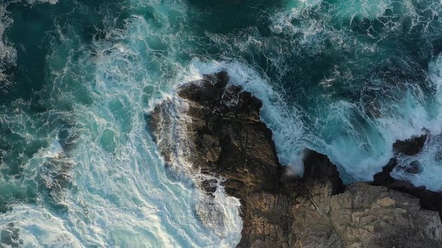 Top aerial drone view of waves crashing on rocky coastline. Blue ocean, wave and impact power. Slowly moving down. 4K.