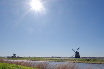 Two windmills in silhouette of a canal under a clear blue sky and the sun