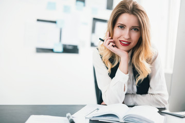 Fototapeta na wymiar young blond beautiful woman manager works in her modern office, holding her hand with pen near face, looks happy, multitasking, work concept