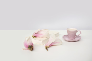 cup of cappuccino on a white table, blooming magnolia buds, a book in a white cover on a delicate pink background, a blank for the designer, an invitation form, a spring mood concept