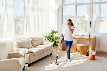 Young woman in white shirt holding vacuum cleaner and talking on cellphone, copy space. Housework,...