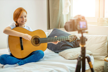 young blogger perform music at camera, red haired little girl play acoustic guitar, self-taught girl present her new song for subscribers