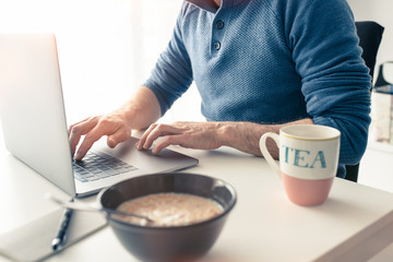 Middle-aged Caucasian man having breakfast while working from home, using his laptop, due to covid-19 coronavirus.