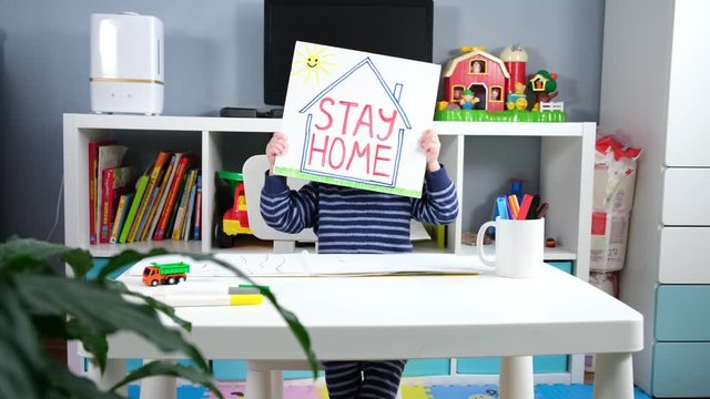 Coronavirus covid-19 slogan stay home. Caucasian boy in home self-isolation holds inscription "stay home" hides his head and looks out from behind paper. Coronavirus don't leave your house on street