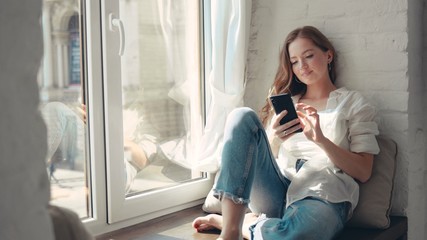 Gorgeous young woman sitting on a windowsill at home and texting on her phone communication female looking message cellphone cheerful smile use internet modern smartphone