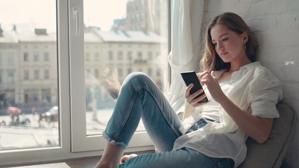 Close up pretty young woman sitting on a windowsill at home and texting on her phone communication female looking message cellphone cheerful smile use internet modern smartphone slow motion