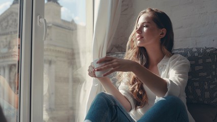 Attractive red hair girl looks out the window at cosy flat itting on a windowsill at home smile drink coffee at sunlight dream holding interior lady lifestyle portrait close up