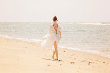 Woman in white dress and mala necklace walking away on the sunny beach by the sea