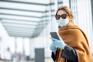 Young woman in face mask, medical gloves and sunglasses with a smart phone at the public transport...