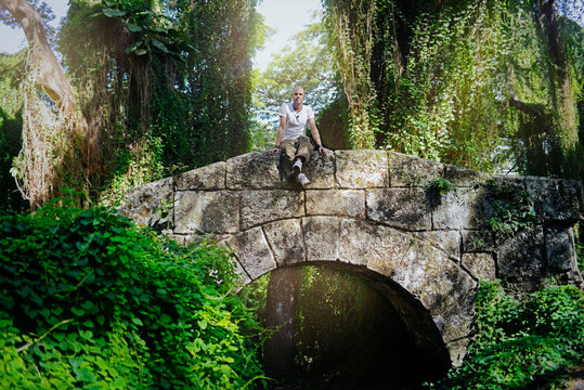 Joven sentado encima de un puente de piedras mohosas en el bosque de La Habana . Pensando al aire libre con anti,is,o, emprendimiento, meditando, tranquilamente, feliz.