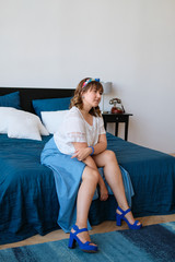 Fototapeta premium A young plus-size woman is sitting on a bed in the bedroom. The smile on her face, the flirtatious look in her eyes. Happiness and beauty in any weight. Girl in a long skirt, high-heeled sandals