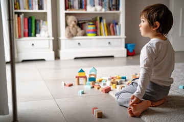 Little sad thoughtful bored toddler boy playing wooden colorful building blocks alone at home...