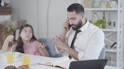 Portrait of busy Middle Eastern father ignoring little daughter eating breakfast. Young businessman talking on the phone while cute girl touching his hand. Lifestyle, multitasking, parenthood. - Powered by Adobe