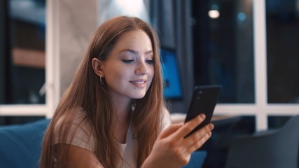 Beautiful young caucasian red hair woman relaxed sitting on sofa browsing on phone and smile at modern apartment Texting sharing messages on social media enjoying feel happy mobile technology evening
