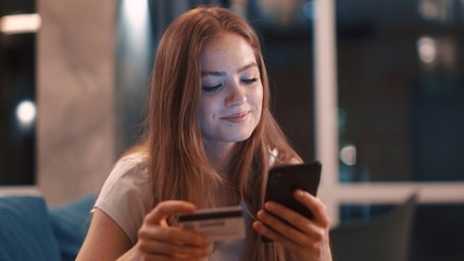 Charming happy girl make online payment holding bank card using modern smartphone or cell phone at...