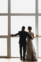 adult bride and groom hug and stand near a large window with their backs to the viewer