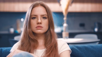 Portrait of serious beautiful red hair woman in white t-shirt sitting in blue coach and looking to camera in modern apartment