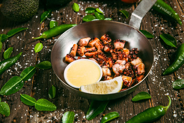 Fried octopus tentacles with oil and lemon in a metal bowl on a wooden background with decorations in the form of green leaves and green peppers