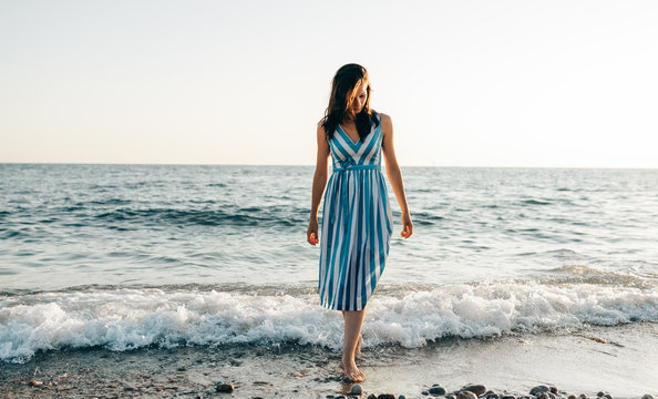 Full body image of beautiful woman walking along the beach and the sea sunset background. Brunette female wearing striped dress has dreamy expression against ocean background. Travel concept.