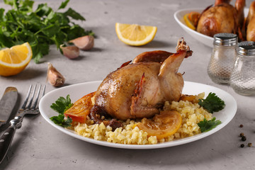 Baked quails with lemon and orange served with bulgur on a white plate on a gray background