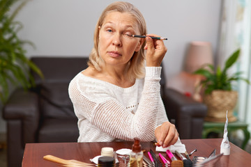 Pretty, aged woman applying black mascara, dye eyelashes using tassel, making, doing professional make.