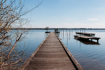 A large blue lake where the blue sky is reflected, with a wooden jetty and a tree. The day is sunny with clouds in the sky.