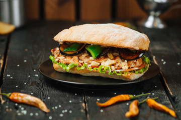 Ciabatta sandwich with chicken, eggplant and sauce on a black plate on a dark wooden table, horizontal, side view