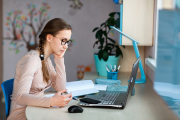 Beautiful young school girl working at home in her room with a laptop and class notes studying in a...