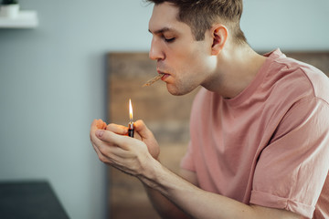 young caucasian man going to smoke cannabis weed at home, he warming hemp medicine with lighter. indoors