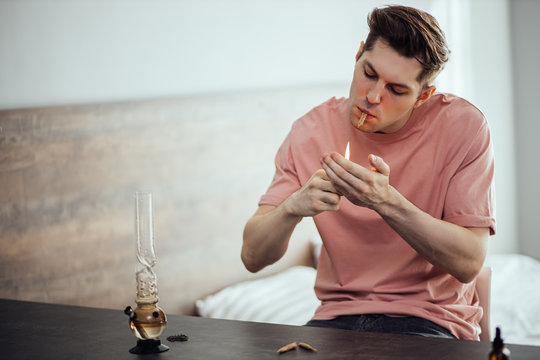 Handsome Caucasian Man Smoking Cannabis Weed At Home In Bedroom. He Use Bong For Making Cannabis, Warm It With Lighter. Marijuana, Drugs Concept