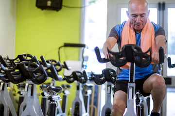 Uomo anziano con maglia blu e asciugamano arancione al collo si allena facendo spinning in una...