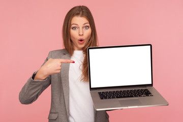 Portrait of amazed pretty brunette businesswoman in elegant suit pointing at mock up display, holding laptop with blank screen for advertising, looking shocked. studio shot isolated on pink background