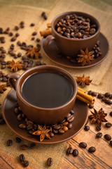 Coffee in dark brown cups and saucers on napkins on a wooden table with spilled coffee beans, star anise, and cinnamon sticks.