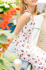 Detail Sunny shot of cheerful young lovely blonde female in white cherry wear dress sitting at table in city cafe, leaning her chin on raised hand and smiling happily to camera