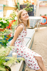 Fototapeta na wymiar Sunny shot of cheerful young lovely blonde female in white cherry wear dress sitting at table in city cafe, leaning her chin on raised hand and smiling happily to camera