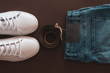 A set of white leather sneakers, belt and blue jeans on brown background. Copy space. Top view.