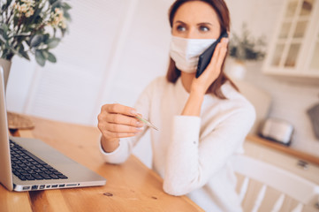 European sick woman in medical mask speaking smartphone, thermometer in hands, working on a laptop, kitchen home quarantine isolation Covid-19 pandemic Corona virus. Distance online work, stay home