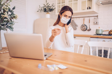 European sick woman in medical mask speaking smartphone, thermometer in hands, working on a laptop, kitchen home quarantine isolation Covid-19 pandemic Corona virus. Distance online work, stay home