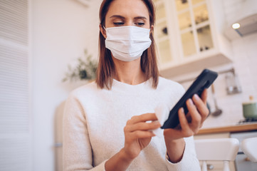 European woman in face protection mask cleaning mobile phone to eliminate germs Covid-19 by hand...